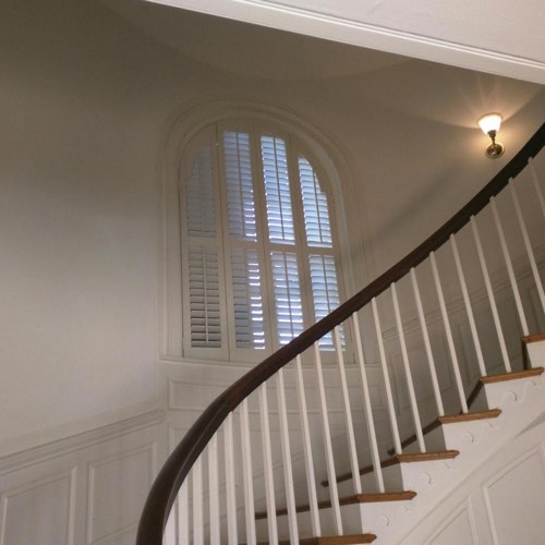 White plantation shutters covering arched window located in curved stairwell.